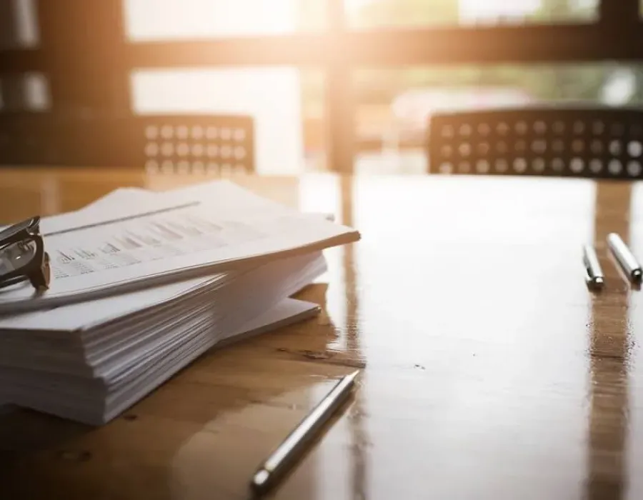 Stack of legal papers on a sunlit desk with glasses rested on top and pens on table nearby